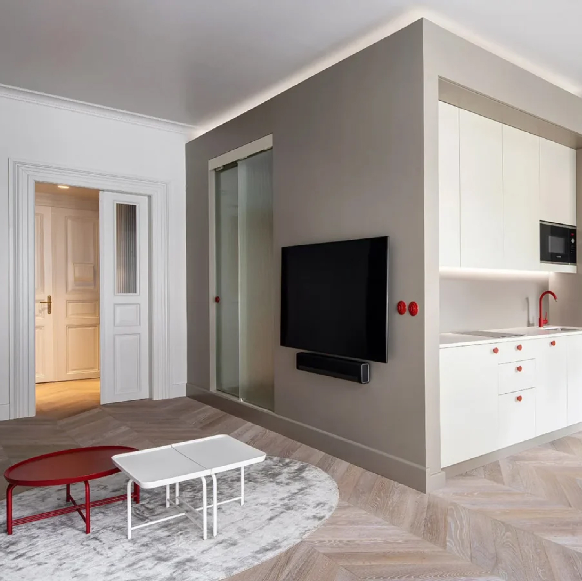 Living room and kitchen organized around a central architectural volume, featuring white cabinetry, herringbone oak flooring, glass partitions, and red accents throughout the interior.