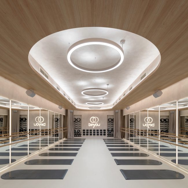 Large yoga studio with mirrored walls, natural oak ceiling details, continuous integrated lighting, circular pendant lights, wooden ballet-style rails, and rows of yoga mats.