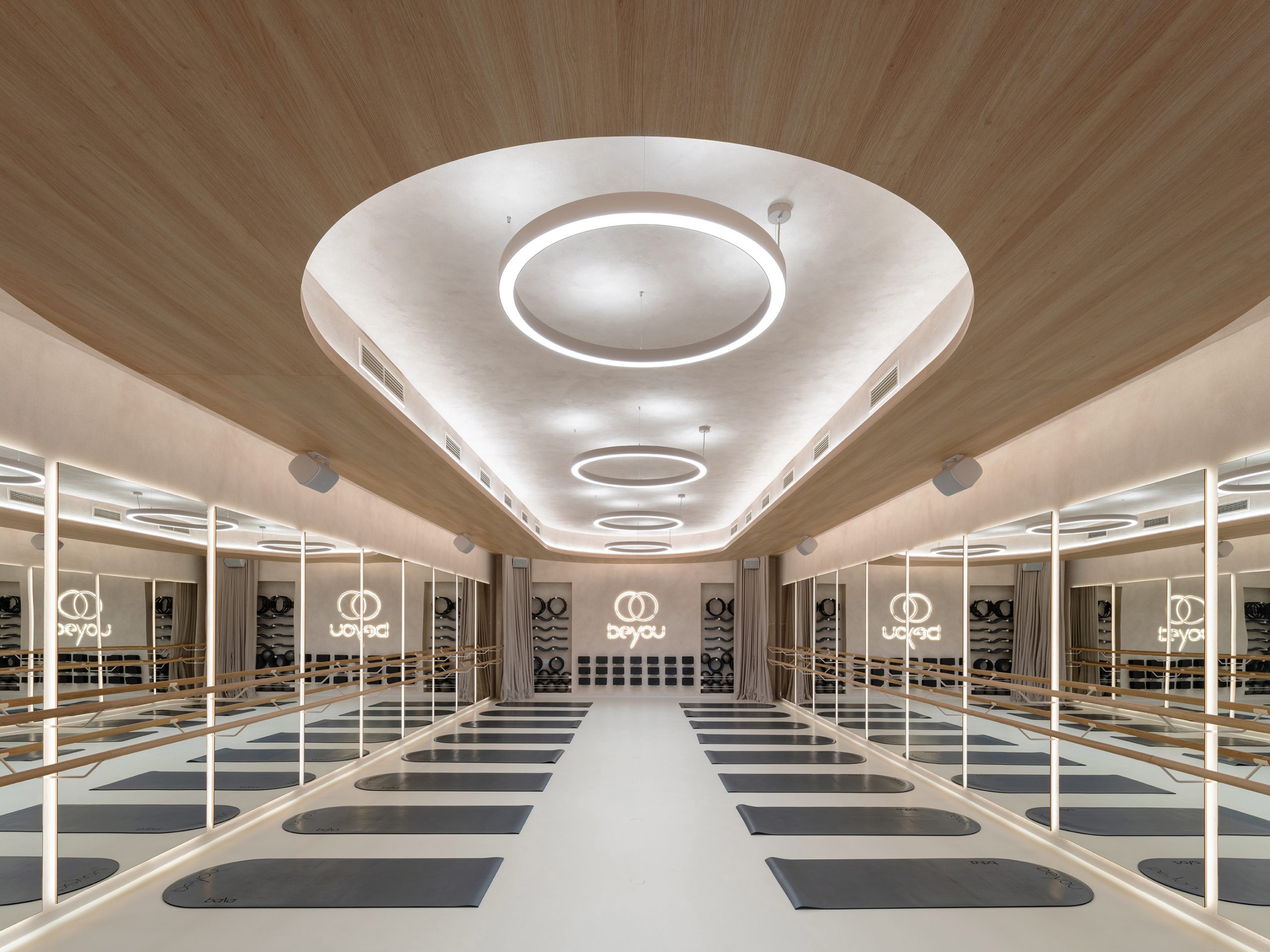 Large yoga studio with mirrored walls, natural oak ceiling details, continuous integrated lighting, circular pendant lights, wooden ballet-style rails, and rows of yoga mats.