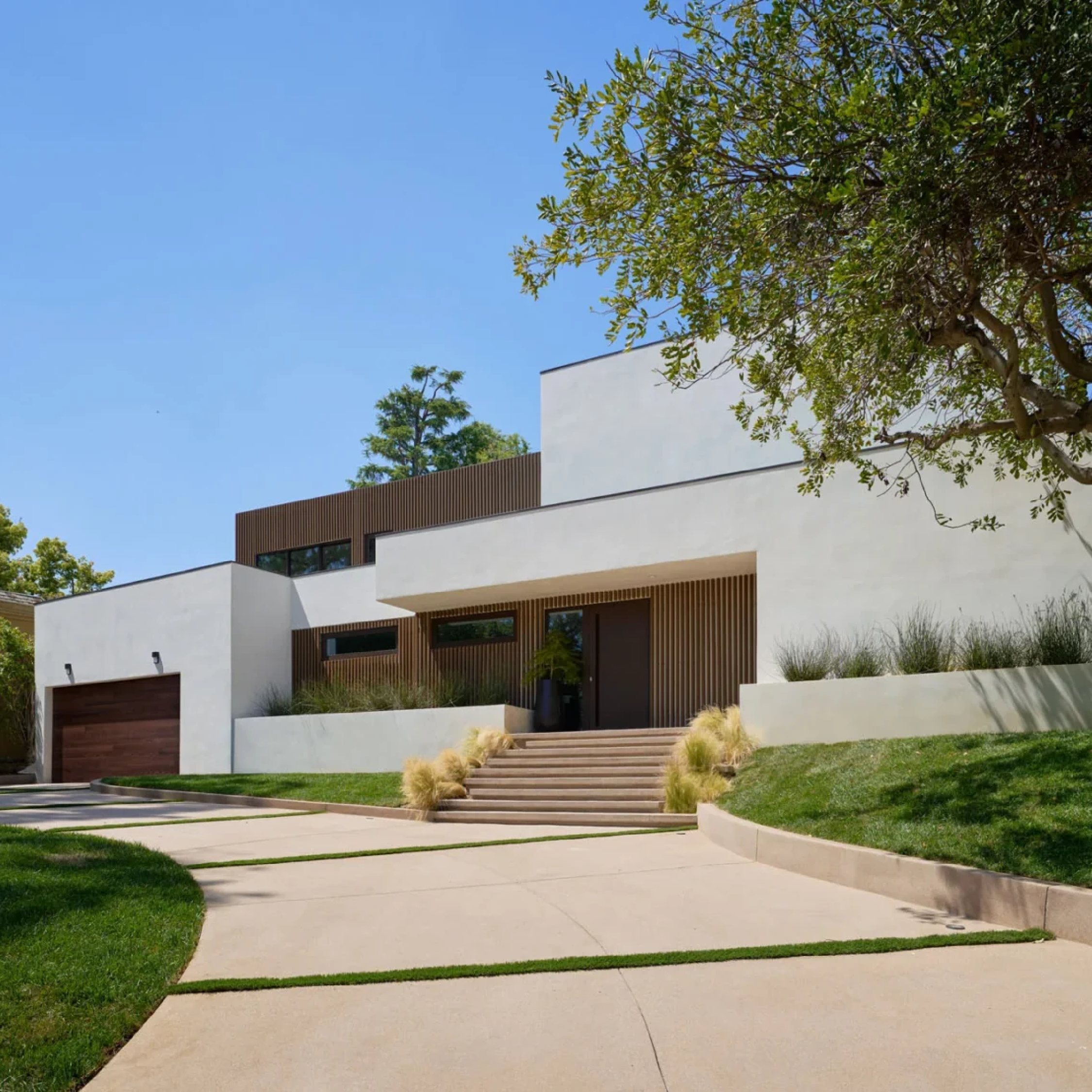 A modern two-story house with clean white walls, flat roof, large windows, wooden accents, and an attached garage with a wooden door. Curved driveway and landscaped yard with trees and ornamental grasses.