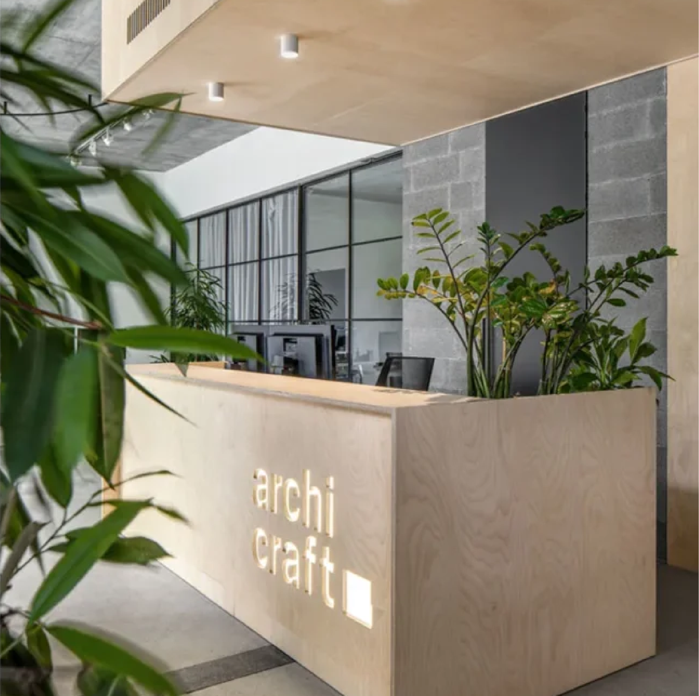 Archicraft architecture studio reception featuring a light wood desk surrounded by plants, with exposed concrete walls and large glass panels overlooking the interior workspace.