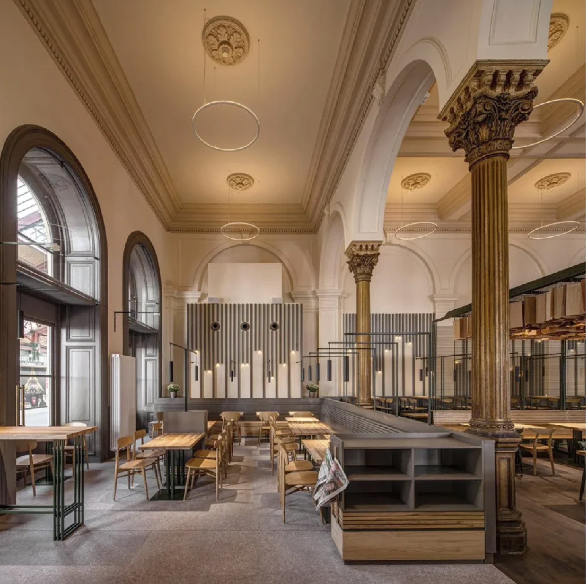 Masaryčka Restaurant by Archicraft, featuring restored ornate columns, arched windows, decorative plaster ceilings, oak dining tables, and contemporary steel partitions within a historic railway station hall.