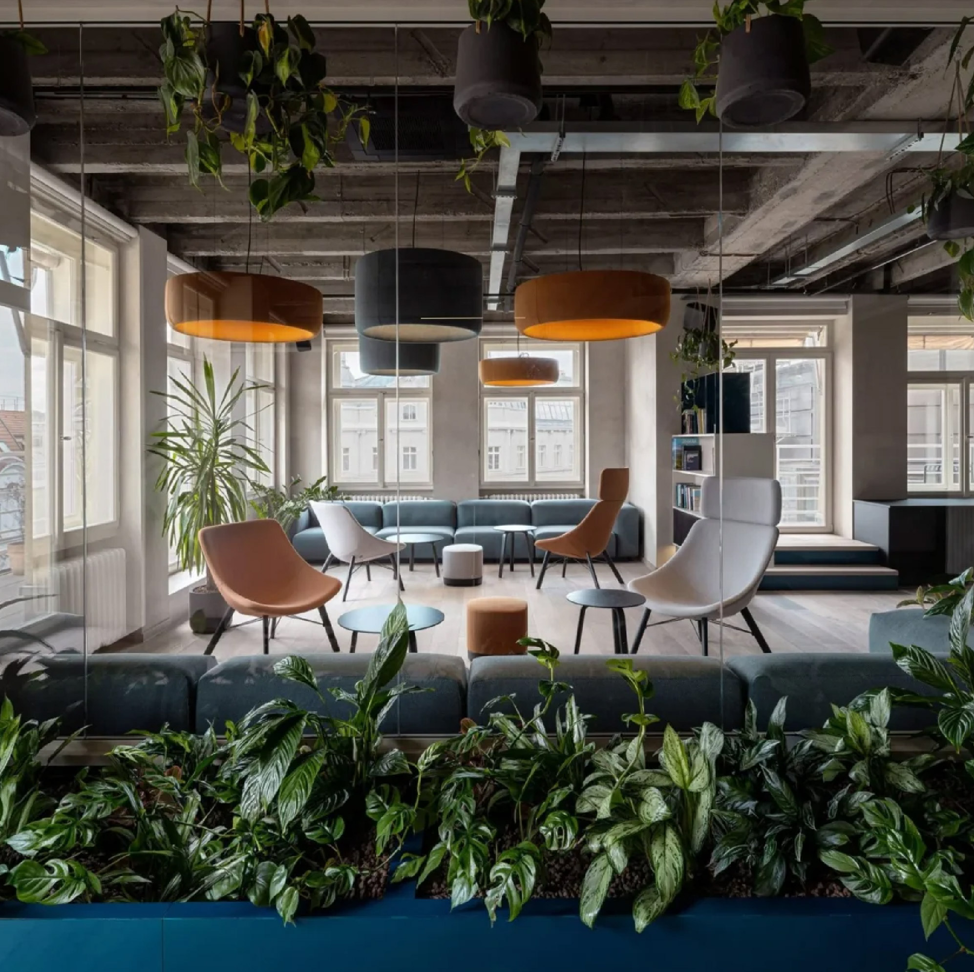 Office That Breathes by Archicraft, featuring an exposed concrete ceiling, blue upholstered seating, cognac and white lounge chairs, warm pendant lighting, and lush greenery throughout the interior.