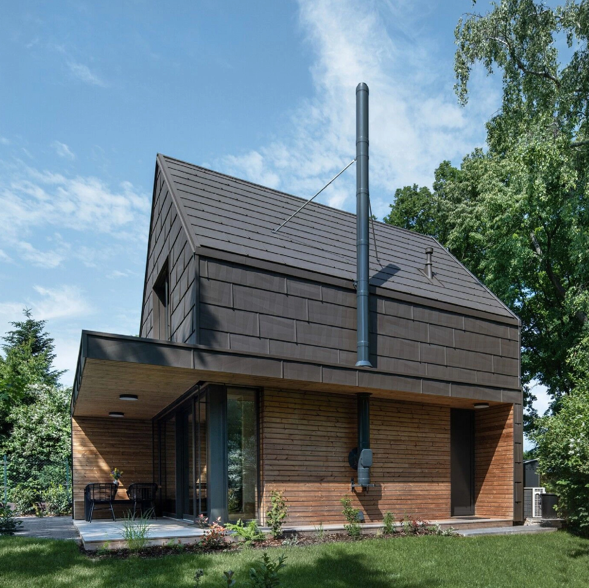 Contemporary cottage by Archicraft featuring dark metal shingle cladding, warm timber siding, a pitched roof, and a covered terrace surrounded by trees and greenery.