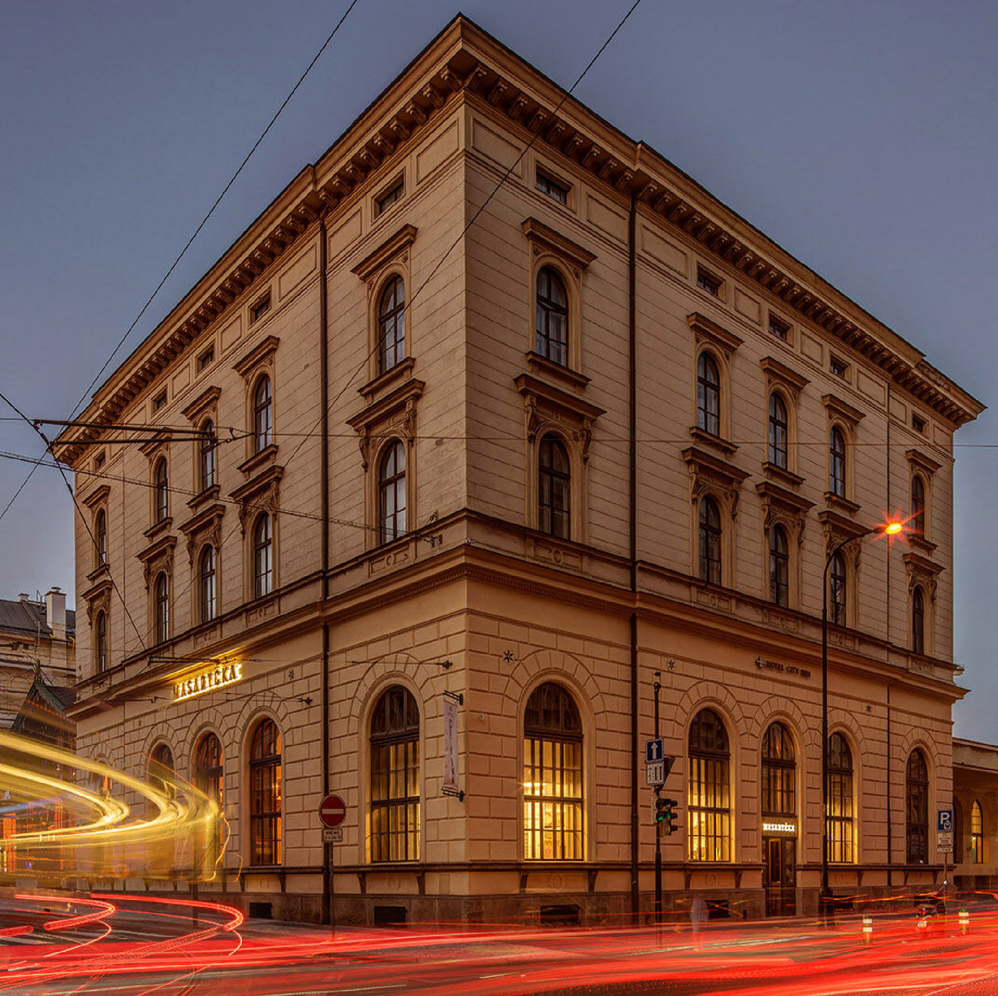 Historic Masaryčka Restaurant in Prague’s Masaryk Railway Station, featuring a classical stone facade, arched windows, illuminated interiors, and passing traffic captured as light trails at dusk.